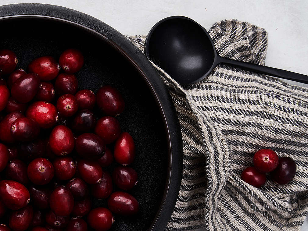 Свежая клюква в черной миске, подробный вид, под ней лежит полосатое кухонное полотенце, а справа рядом с миской лежит черная ложка