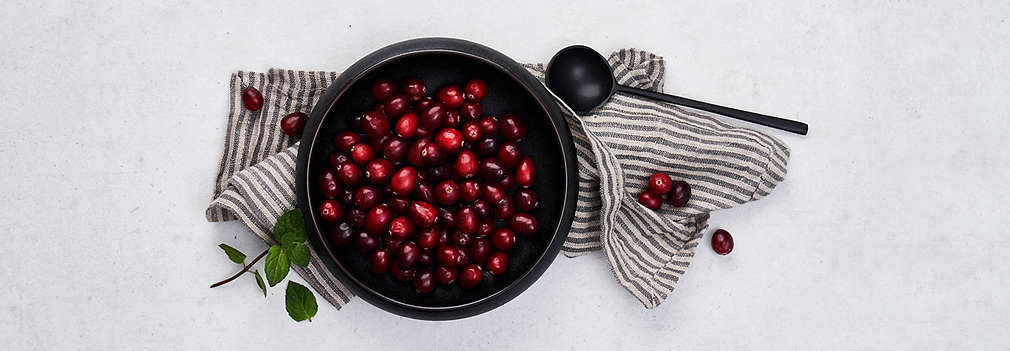 Frische Moosbeeren in einer schwarzen Schüssel, darunter liegt ein gestreiftes Geschirrtuch, rechts daneben liegt ein schwarzer Löffel