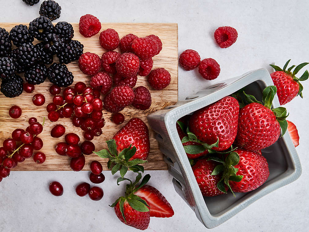 Verschiedene frische Beeren auf einem Holzbrett und Erdbeeren in einer grauen Dose