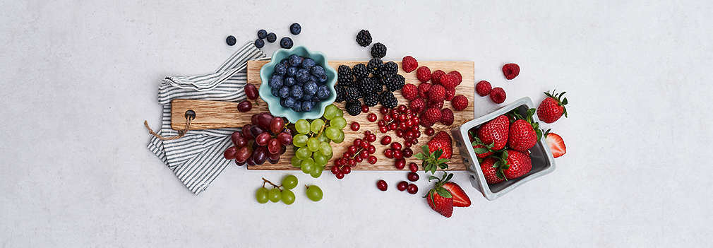 Verschiedene frische Beeren auf einem Holzbrett, links daneben Erdbeeren in einer grauen Dose, rechts liegt ein gestreiftes Geschirrtuch unter dem Brett