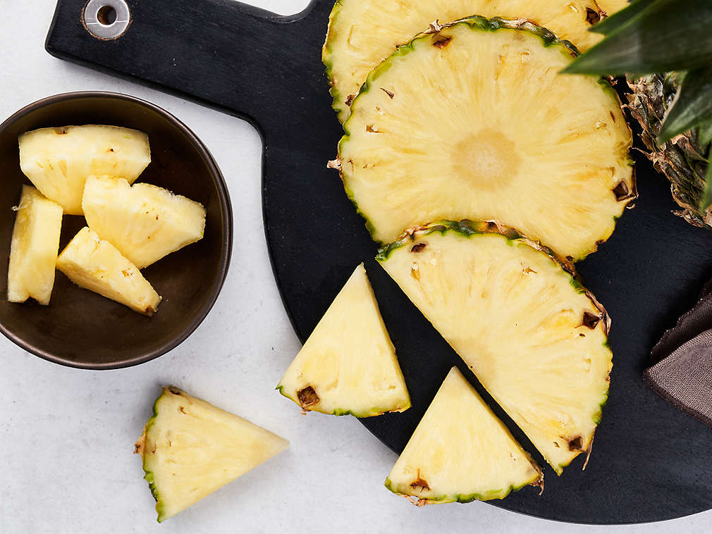Ananas proaspăt tăiat în felii și în bucăți pe un tocător negru și într-un castron mic maro alături