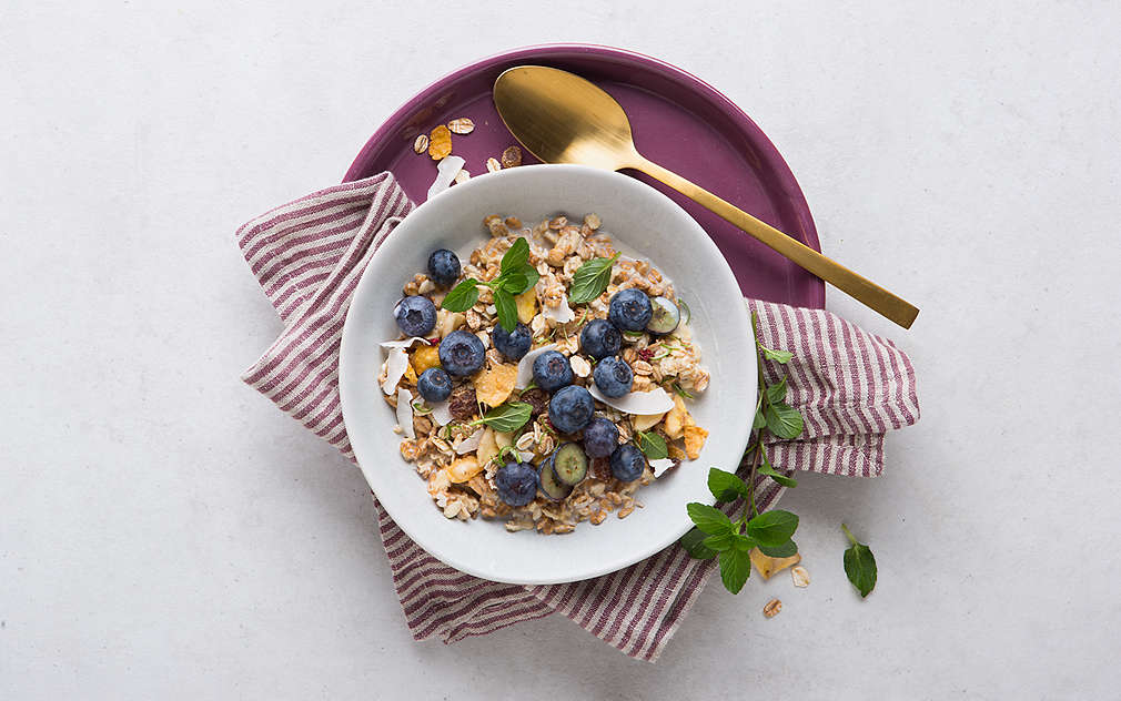 Misa s müsli, ozdobená čučoriedkami a lístkami mäty. Vedľa toho je zlatá lyžica. Pod tým je podnos černicovej farby a červeno-biela pásikavá utierka na riad.