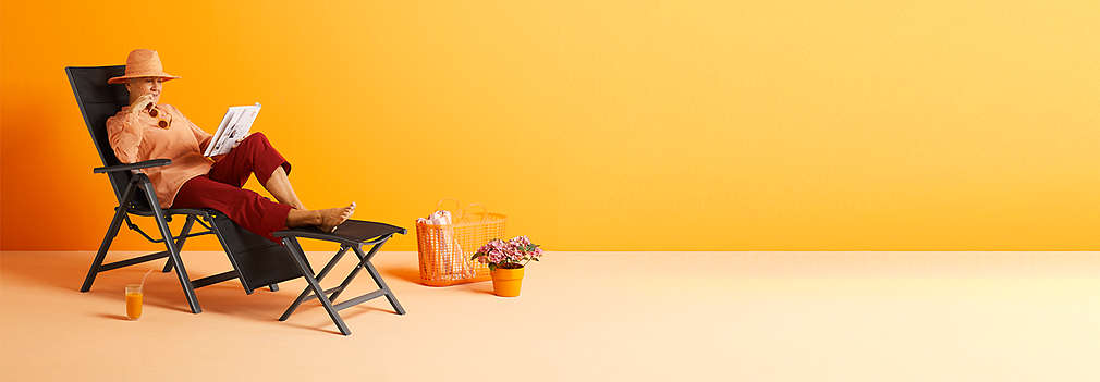 A woman lies on a sun lounger and reads something. Next to it is some decoration. The scenery is placed on an orange background.