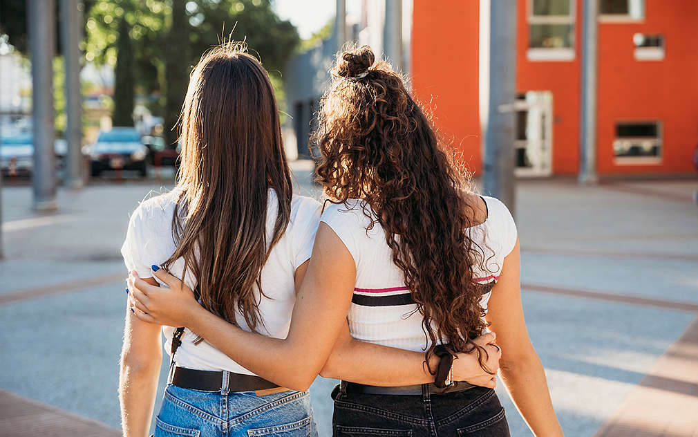 Две жени с дълги тъмни коси, снимани откъм гърба. Те се прегръщат.
