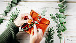 Na obrázku je štvorcový oranžový balíček, na ktorom je položený výhonok eukalyptu. Dve ruky ho pripevňujú na balíček.