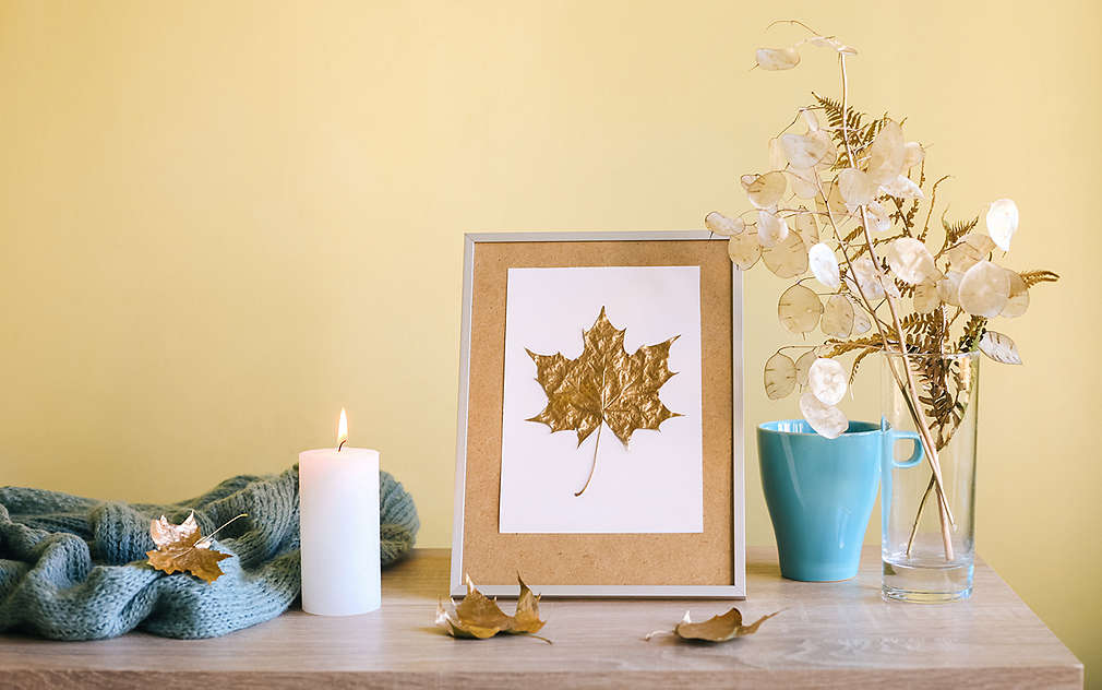 Na obrázku je: zarámovaný obrázok so sivým rámom, bielym pozadím a javorovým listom so zlatým povrchom. Obrázok je položený na stole. Na stole je okrem toho: tyrkysová hrejivá deka, zapálená biela sviečka, vysoká tyrkysová šálka, sklená váza so sušenými kvetmi, tri javorové listy.