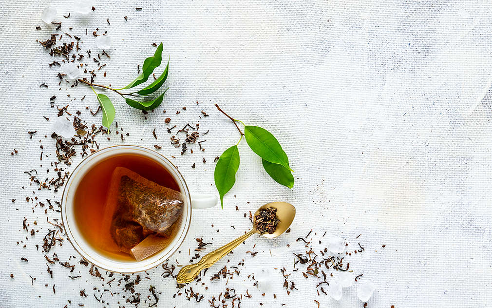 Na zdjęciu: filiżanka z torebką herbaty, łyżeczka i luźne liście herbaty.