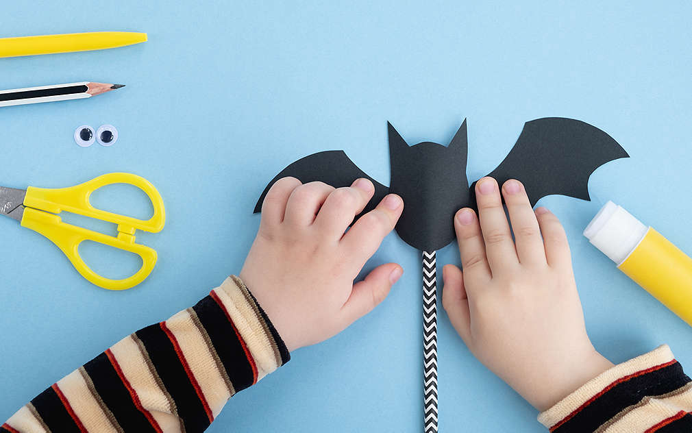 Plava podloga, na njoj žuti pribor za izradu (olovka, škare, ljepilo) i crno-bijela olovka, u sredini slike dvije dječje ruke izrađuju crnog šišmiša od papira koji je pričvršćen za štapić.