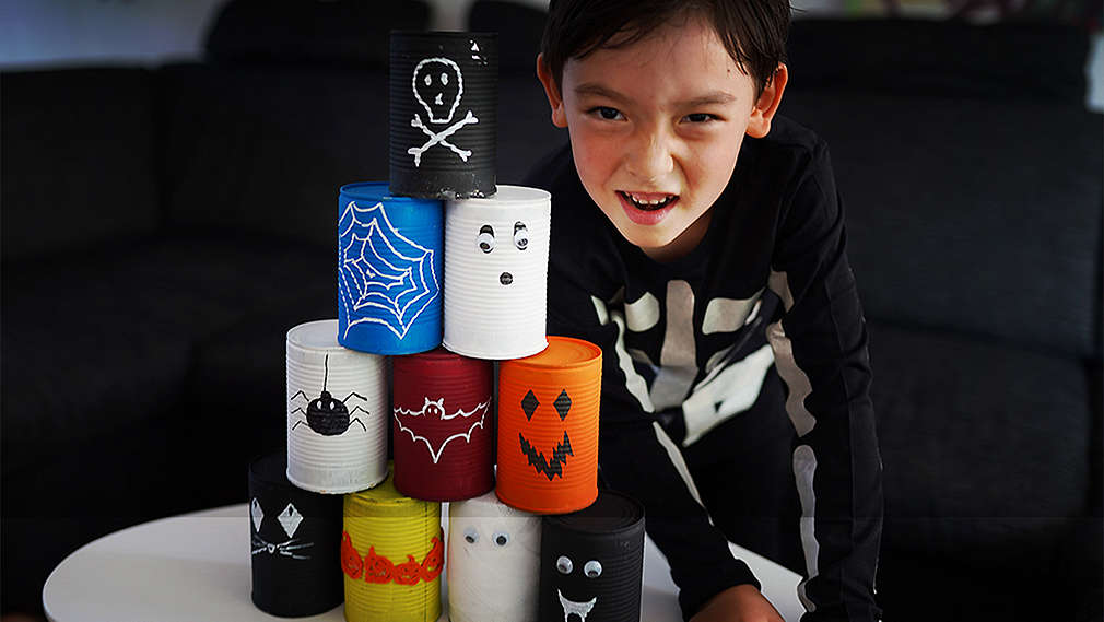 Un băiat îmbrăcat în costum de Halloween privește cu fața la cameră un turn de conserve pictate.