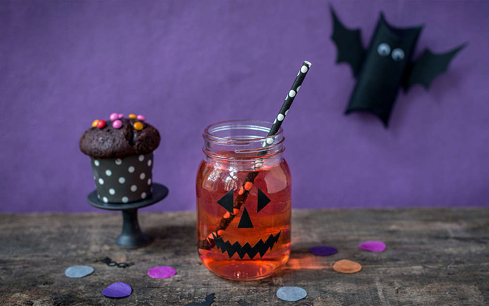 Na stole stoi szklanka z naklejoną twarzą halloweenowego upiora. W szklance jest czerwony płyn i czarno-biała słomka. Po lewej stronie jest czekoladowa muffinka owinięta w czarno-biały papier.