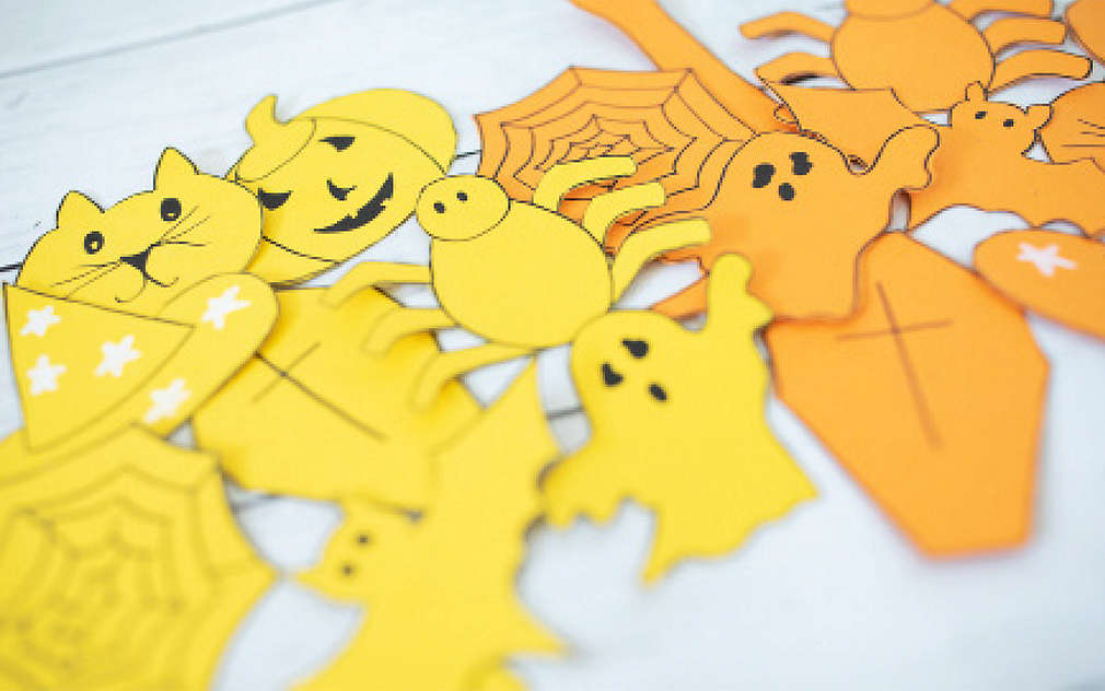 Na jasnym stole leżą wycięte z papieru żółte i pomarańczowe postaci halloweenowe.