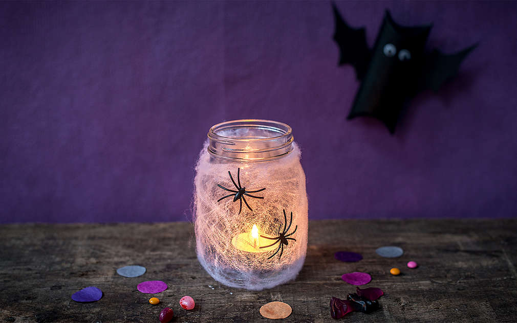 Słoik ze świeczką podgrzewaczem jest z zewnątrz ozdobiony sztuczną pajęczyną i czarnymi pająkami.