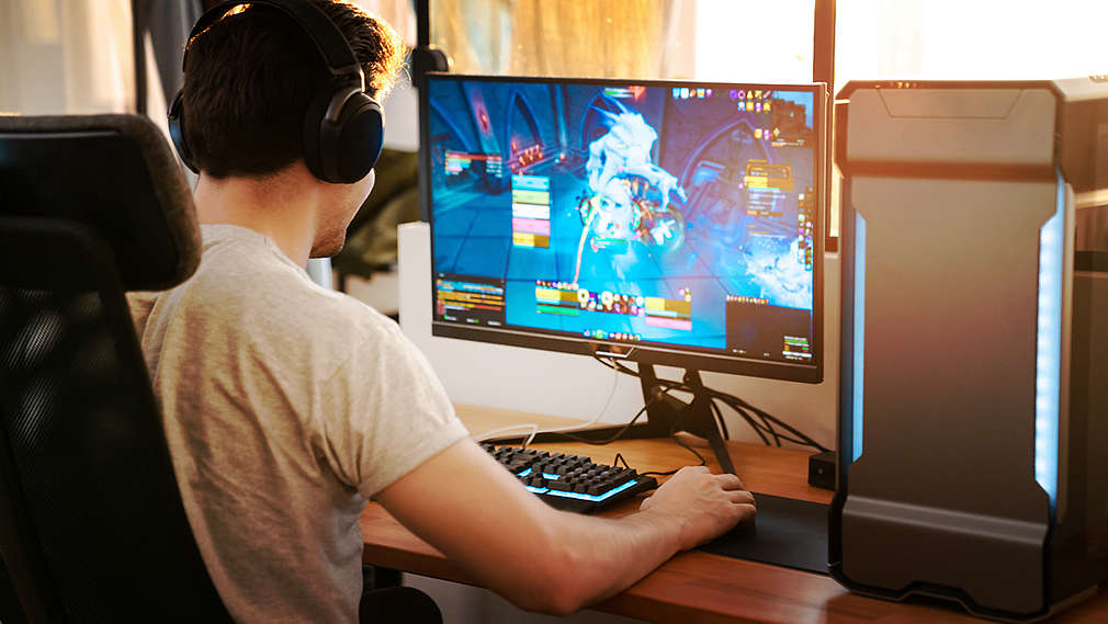 Вижда се млад мъж, който седи пред компютъра и играе компютърна игра на голям екран.