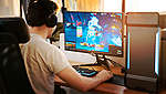 Se poate vedea un tânăr care stă în fața unui calculator și se joacă un joc pe calculator în fața unui monitor mare.