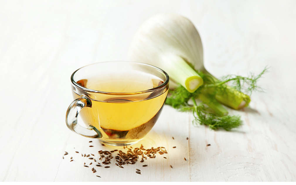 Šálek fenyklového čaje. Před ním je několik fenyklových semínek, za ním čerstvá fenyklová cibulka.
