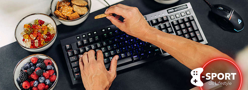 Eine Person greift über eine Gaming-Tastatur hinweg nach einem Cracker, daneben eine Gaming-Maus sowie Schälchen mit Beeren und Gummibärchen