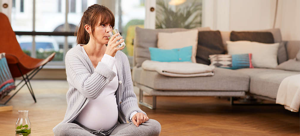 Schwangere sitzt im Schneidersitz und trinkt ein Glas Wasser