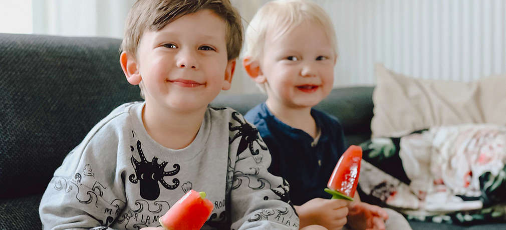 Zwei Kinder sitzen lächelnd auf dem Sofa und halten selbst gemachtes Eis in der Hand