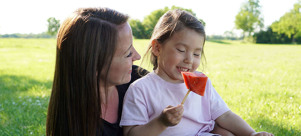 Mutter und Tochter sitzen auf einer Picknickdecke und essen Melone am Stiel