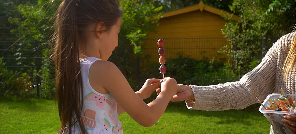 Ein Kind erhält von der Mutter als Sommersnack einen Spieß mit gefrorenen Trauben im Garten