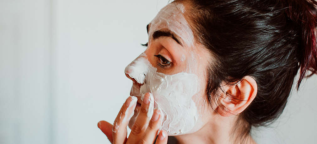 Eine junge Frau mit zusammengebundenen Haaren in ihrem Badezimmer, die eine cremige Maske auf ihr Gesicht aufträgt 