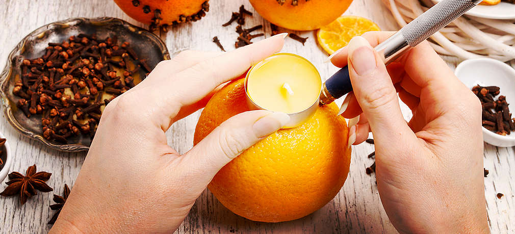 Eine Orange wird von zwei weiblichen Händen mithilfe eines Messers zu einer Kerze vorbereitet. Links daneben befinden sich Nelken auf dem Tisch.