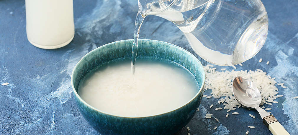 Blauer Untergrund, darauf milchig-weiße Plastikbehälter, ein Löffel mit weißem Griff, Reiskörner und eine türkise Schale mit Reiswasser, in die gerade Wasser aus einem Krug gegossen wird, Hand im Anschnitt.