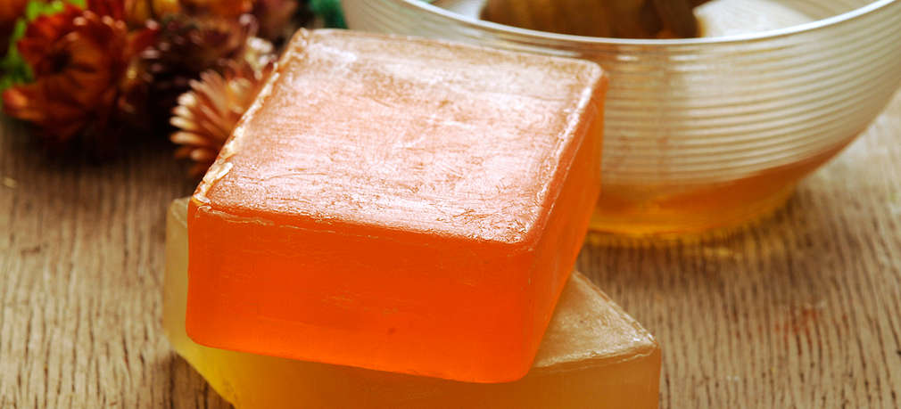 Zwei Stück handgemachte Seife – oben in oranger, unten in gelber Farbe, die übereinander gestapelt auf Untergrund aus Holz liegen. Im Hintergrund ist eine Glasschale mit Honig.