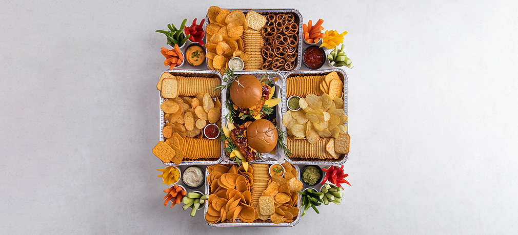 Aus der Vogelperspektive ist ein Snackstadion zu sehen, das aus mehreren Grillschalen besteht, das in der Mitte mit Guacamole, außerdem mit Gemüsesticks, Crackern und Chicken Wings befüllt ist. Rund um das Stadion sind außerdem Wraps, Sandwiches und Dips platziert.