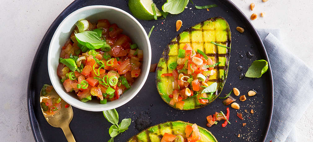 Zu sehen: heller Untergrund, helles Geschirrtuch, darauf schwarzer flacher Teller. Auf dem Teller: zwei gegrillte Avocadohälften belegt mit Tomatensalsa, Schälchen mit Tomatensalsa, Basilikumblätter, goldener Löffel