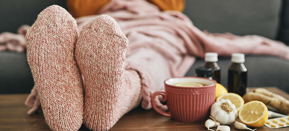 Zu sehen ist eine Person, die auf dem Sofa sitzt und ihre Füße auf dem Tisch aufgelegt hat. Auf dem Tisch sieht man eine Tasse, Knoblauch, aufgeschnittene Zitronen, Tabletten und kleine Medizinflaschen.