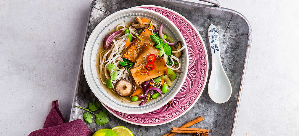 Asiatische Suppenschüssel auf einem grauen Emailgeschirr, gefüllt mit einer Pho-Suppe mit Pak Choi, Nudeln und Tofuscheiben. Auf dem Tablett sind außerdem ein weinrotes Geschirrtuch, eine aufgeschnittene Limette und zwei Zimtstangen