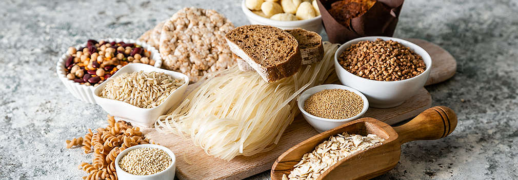 Auf einem Holztisch liegen sowohl ein grauer kleiner Teller mit zwei mit Frischkäse, Schinken und Cherrytomaten belegten Brotscheiben als auch eine Schieferplatte mit Geschirrtuch, Messer und einem angeschnittenem Brot in Kastenform. 