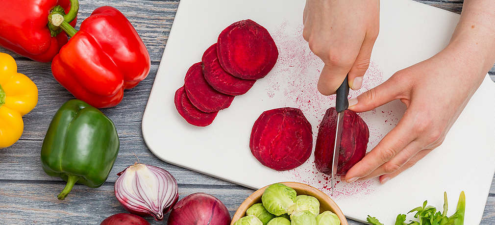 Zwei Hände die rote Beete auf einem Brett schneiden, daneben liegen Zwiebeln, Paprika, Kräuter und Rosenkohl in einer Schüssel