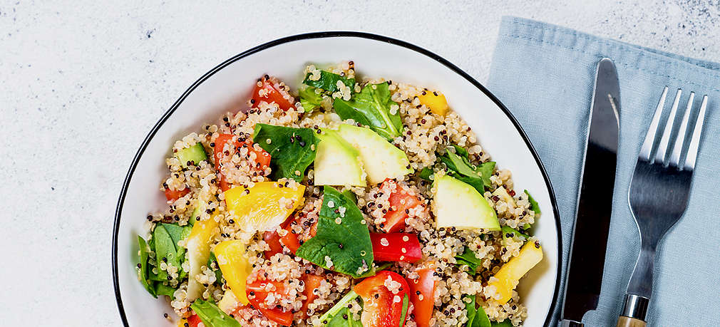 Zu sehen ist eine weiße Schale mit blauem Rand, gefüllt mit einem Gericht aus Quinoa, Paprika, Tomaten, Spinat und Avocado. Links von der Schüssel liegt eine Gabel, rechts davon eine halbe Avocado, zwei Spinatblätter und eine Cocktailtomate. In der linken Ecke des Bildes sieht man einen Teil eines weiß-grau karierten Tischtuchs