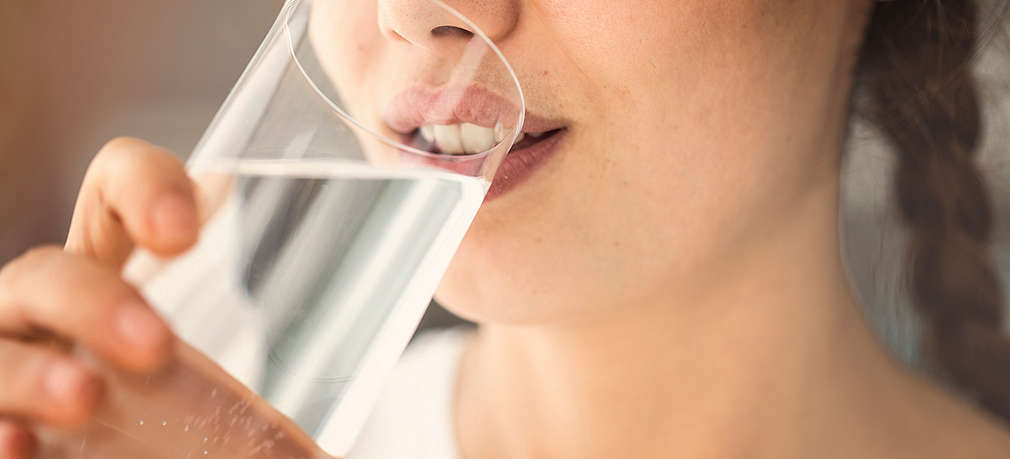 Frau, von der man im Anschnitt Gesicht und Hals sehen kann, trinkt aus einem Wasserglas