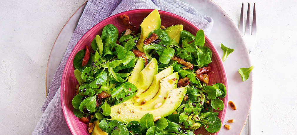 Eine von oben fotografierte pinke Schüssel, in der sich neben Feldsalat auch Avocadoscheiben, Speck und Pinienkerne befinden