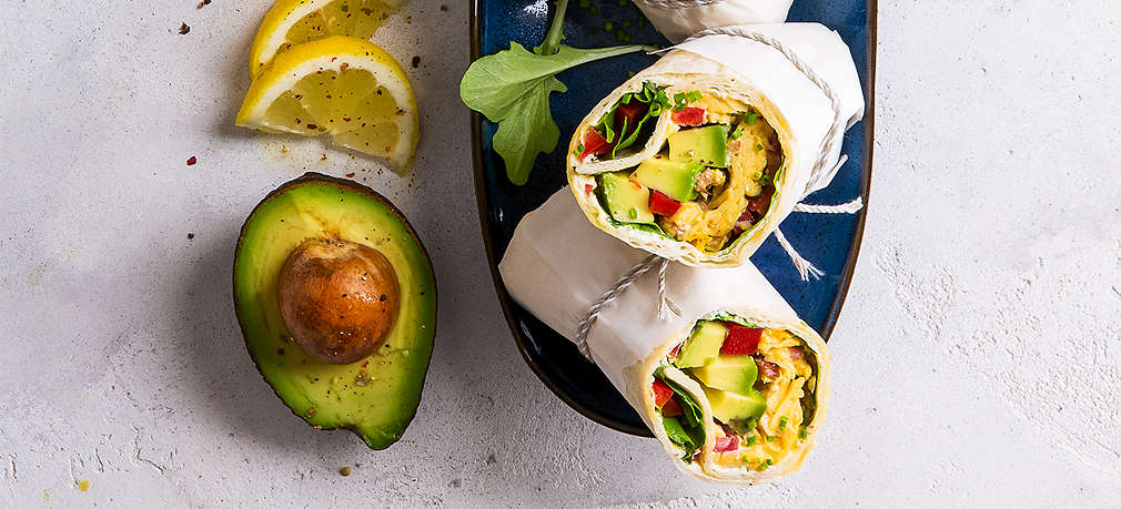 Hellgrauer Untergrund, darauf Pflücksalatblätter, Zitronenscheiben, eine halbe Avocado und ein länglicher blauer Teller mit gefüllten Wraps