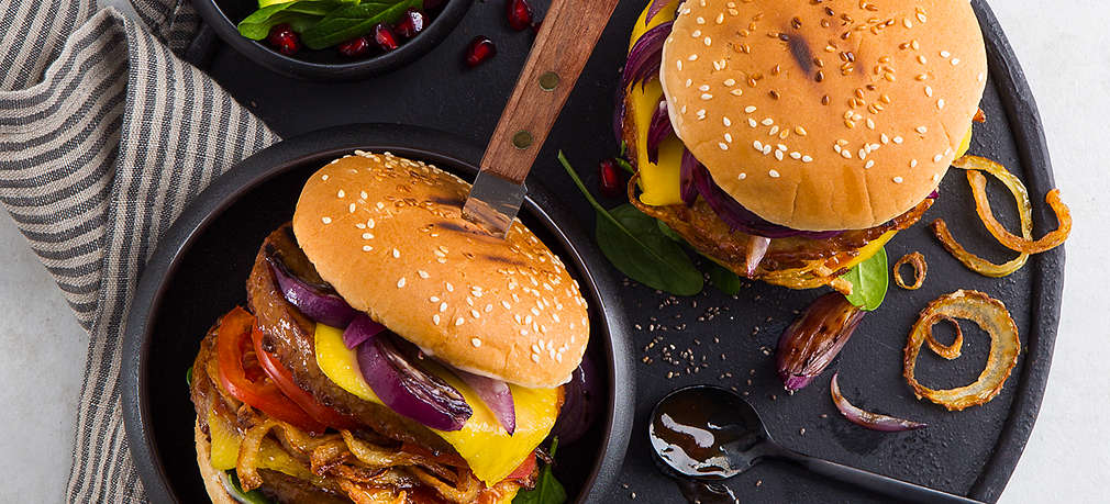 Zwei vegane Burger auf einem schwarzen, runden Teller. Die Burger sind belegt mit gegrilltem Gemüse, unter anderem Zwiebeln, Tomaten und Spinat, sowie einem veganen Burger-Patty und veganem Käse. Daneben steht in einer kleinen Schüssel ein Salat aus Avocado und Granatapfelkernen. 