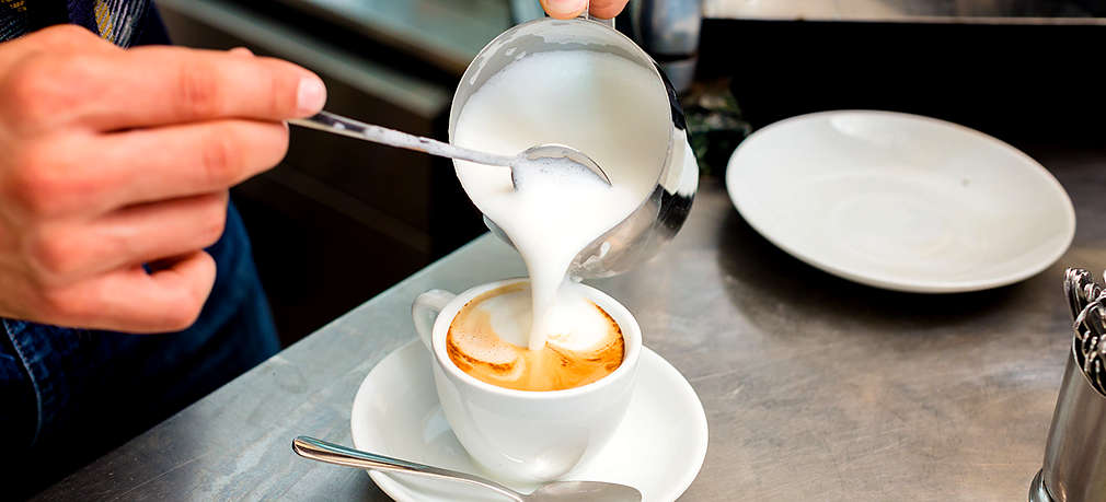 Aufgeschäumte Milch wird aus einem kleinen Kännchen mit Hilfe eines Teelöffels in eine Tasse mit Kaffee gegeben