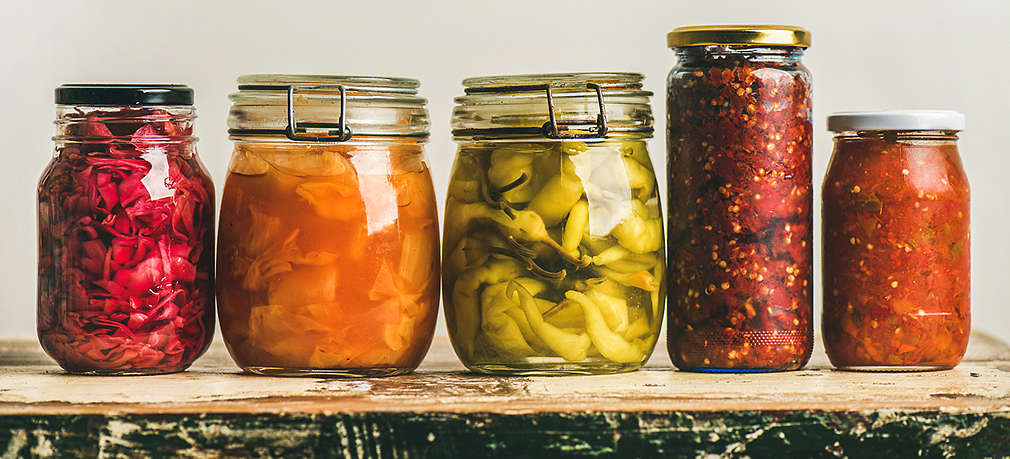 Auf einer Holzfläche stehen fünf Einmachgläser mit unterschiedlichen Sorten fermentiertem Gemüse