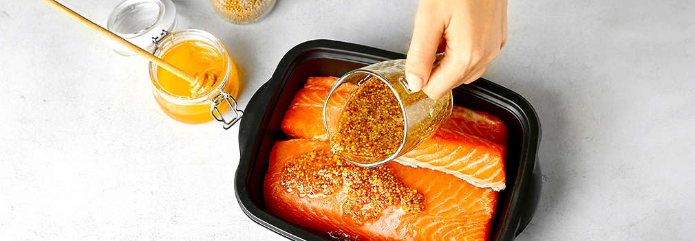 Frischer Lachs wird in einer schwarzen Auflaufform mit Honig-Senf-Marinade mariniert. Die Zutaten stehen ebenfalls auf dem Tisch.