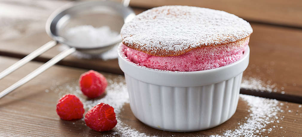 Himbeersoufflé in einer kleinen weißen Backform mit Puderzucker garniert, daneben frische Himbeeren