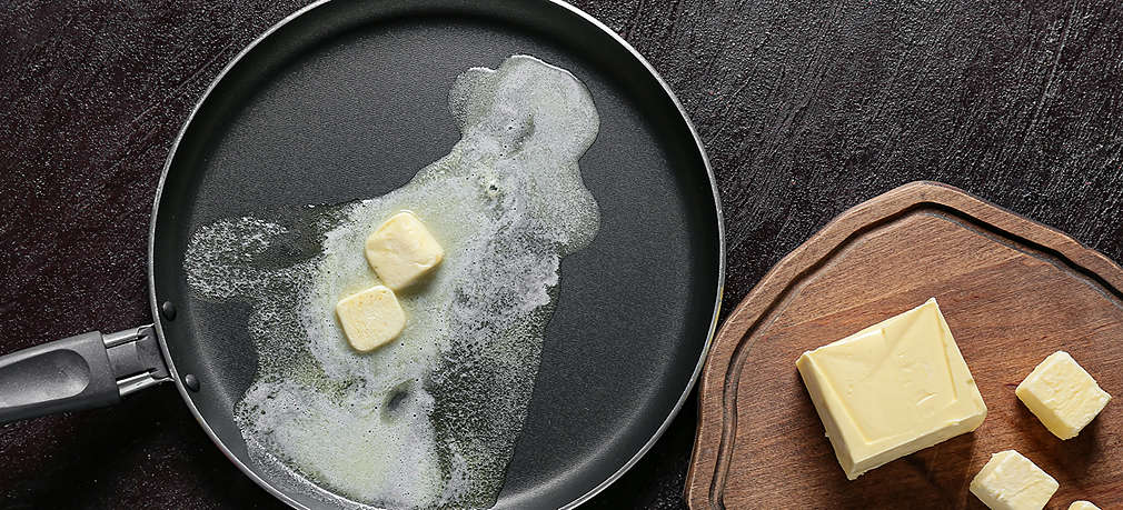 Butter wird in einer Pfanne zerlassen, daneben sind auf einem Holzbrett Butterwürfel zu sehen