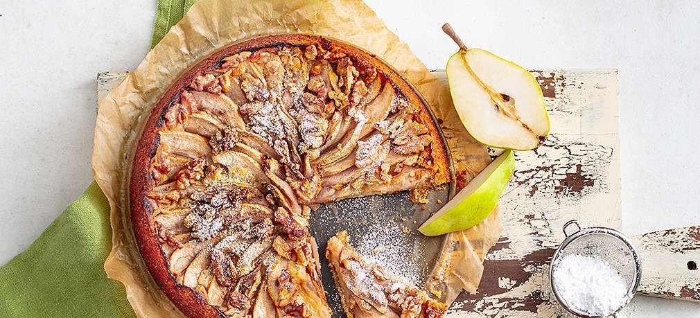 Grauer Untergrund, darauf ein hell lackiertes Holzbrettchen und ein grünes Geschirrtuch, darauf eine runde Backform mit angeschnittenem Kuchen, der mit Birnenscheiben belegt ist. Außerdem im Bild: Halbierte Birne, Kuchenstück, kleines Sieb mit Puderzucker, Schälchen mit Apfelmus.