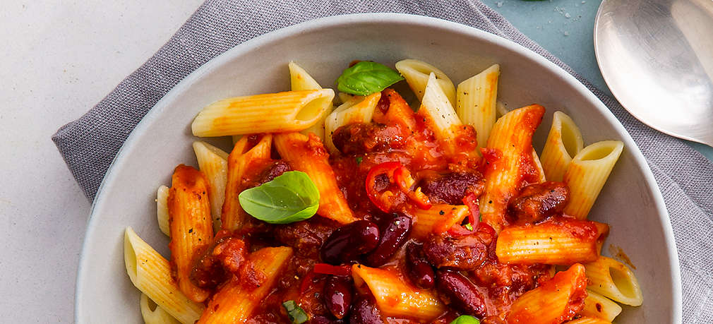 Ein tiefer hellgrauer Teller steht auf hellgrauem Untergrund und einem grauen Geschirrtuch, darin befindet sich eine große Portion Penne mit roter Soße aus Tomaten, Chili und Kindeybohnen, als Garnitur sind einige Blättchen Basilikum auf den Nudeln.