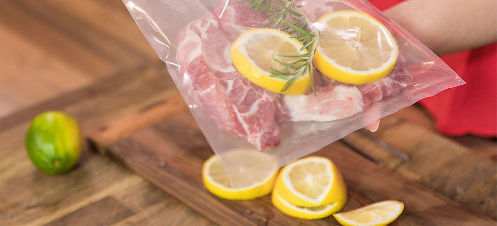 Oberkörper einer Frau im Anschnitt, zwei Hände halten einen Plastikbeutel, darin befindet sich ein Stück Fleisch, Zitronenscheiben und ein Zweig Kräuter, welches für das Sous-vide-Garen vorbereitet wird. Die Frau arbeitet über einer Arbeitsfläche aus dunklem Holz