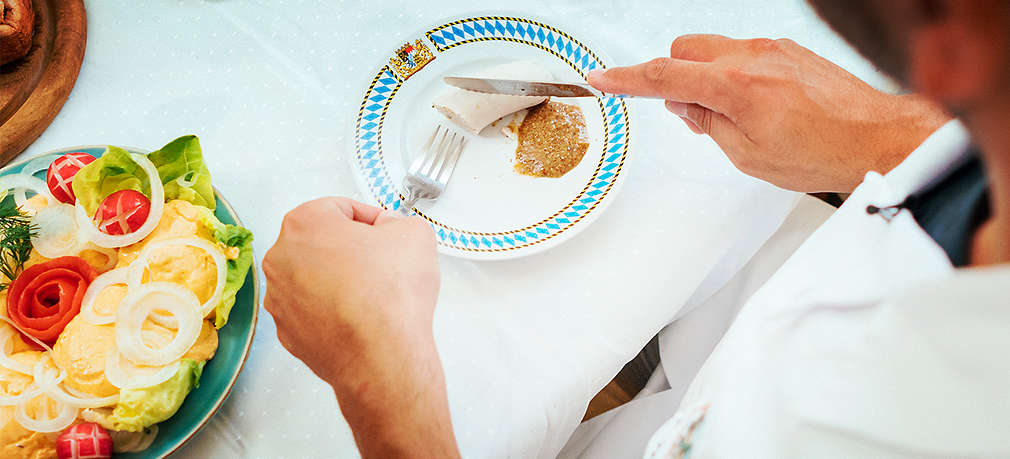 Ein Mann schneidet mit Messer und Gabel eine bereits halb aufgegessene Weißwurst auf einem Teller mit traditionellen bayerischen Mustern an; ebenfalls auf dem Teller ist ein Klecks süßer Senf; daneben steht ein weiterer Teller mit angerichtetem bayerischem Obazda mit Zwiebeln, Radieschen und Salat