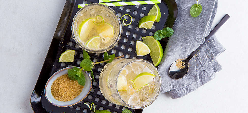 Zwei Cocktailgläser, gefüllt mit Eiswürfeln, Limetten, Minze und Limonade auf einem schwarzen Tablett, darunter ein blaugraues Geschirrtuch, auf dem ein langer Cocktaillöffel liegt. Auf dem Tablett befinden sich außerdem Limettenscheiben, frische Minze, zwei grün-weiße Strohhalme und ein Schälchen mit Rohrzucker