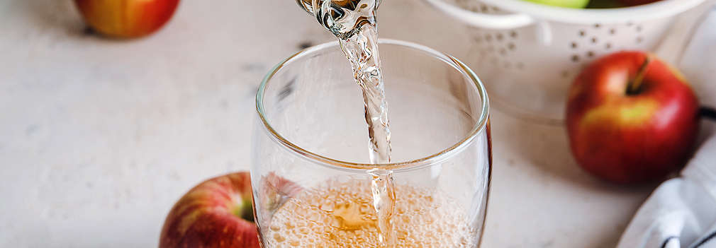 Cidre wird in ein Glas geleert, daneben liegen Äpfel auf dem Tisch sowie in einem weißen Sieb